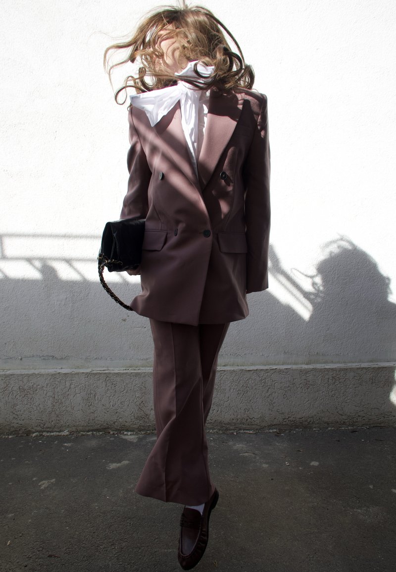 Femeie îmbrăcată în costum de culoarea lavandei și bluză albă sare cu părul acoperindu-i fața, ținând o geantă neagră în fața unui perete alb cu umbre.