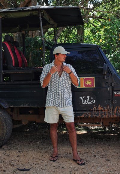 Camisa de manga corta, azul y blanca con un diseño estampado, pantalones cortos ligeros y chanclas, de pie al lado de un vehículo todoterreno negro con asientos con acentos rojos.