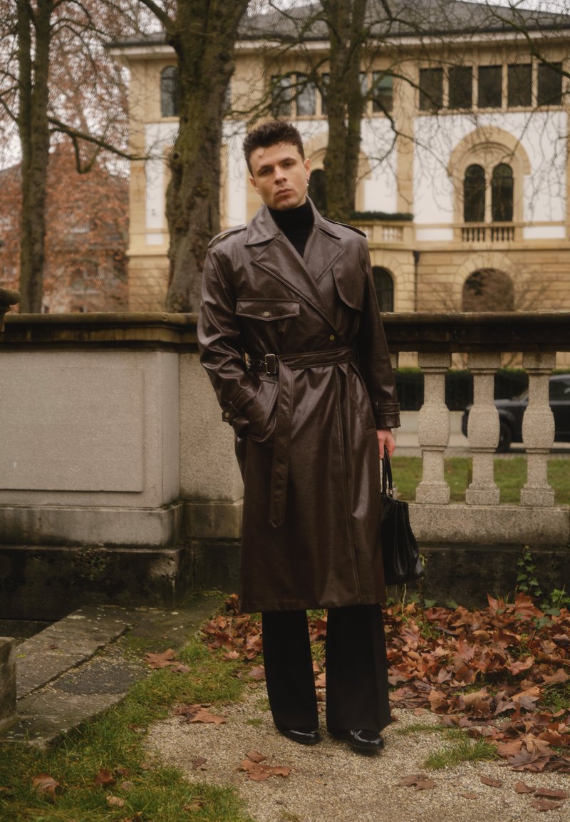 Lungo trench in pelle marrone con cintura in vita, indossato sopra un dolcevita nero e pantaloni neri a gamba larga, abbinato a una borsa nera.