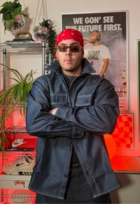 Cămașă din denim cu guler mare, tonuri de albastru și cusături albe. Accesorizată cu o bandană roșie și ochelari de soare. Fundalul prezintă plante și postere.