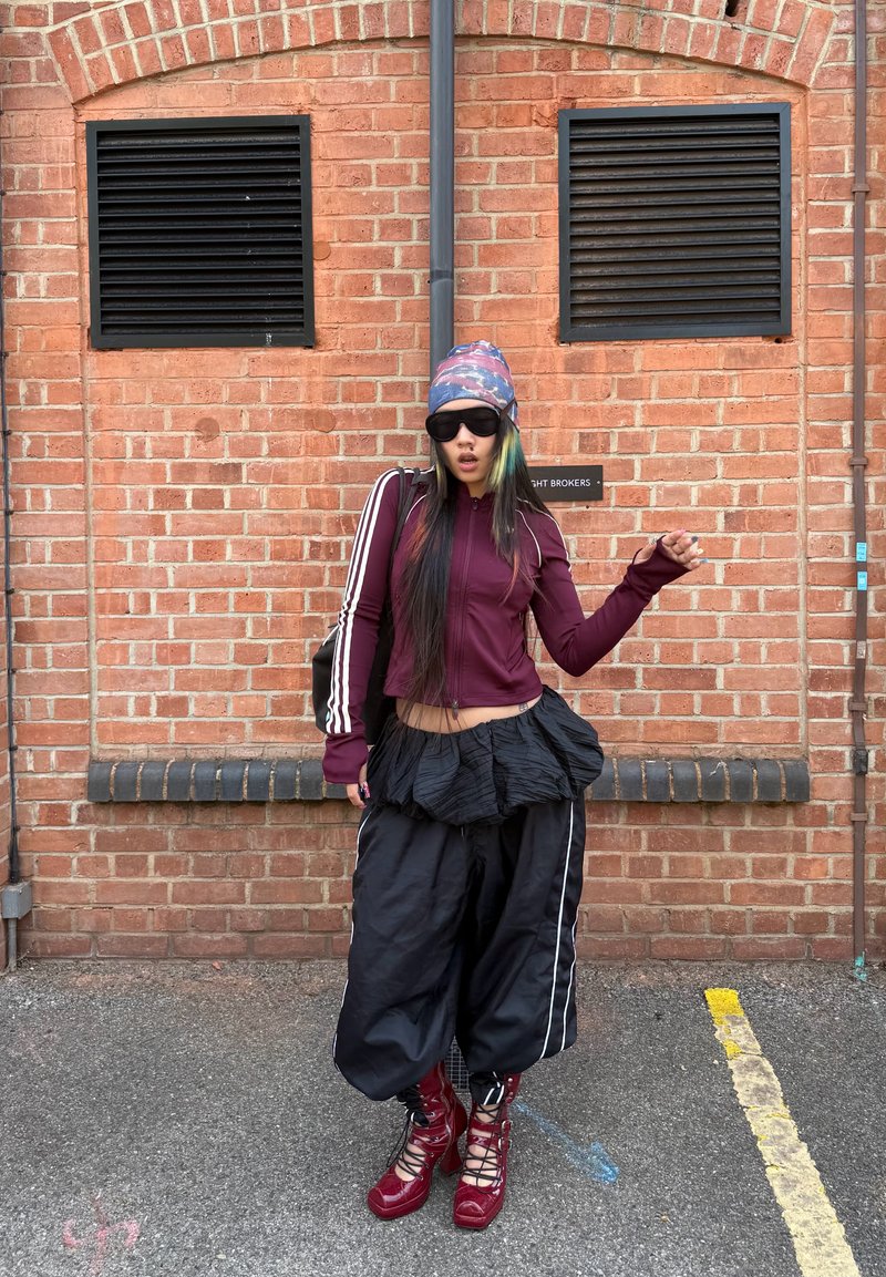 Jeune personne portant une veste bordeaux, un pantalon noir ample, des bottes violettes, des lunettes de soleil et un bonnet multicolore, debout contre un mur en briques rouges.