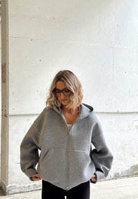 Kobieta o falowanych blond włosach, nosząca duże okulary i szarą bluzę z zamkiem, stojąca wewnątrz pomieszczenia na tle białej, teksturowanej ściany.