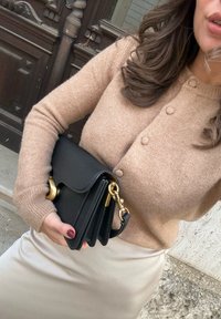 Bežani pleteni kardigan s detaljima na gumbima, držan pored male crne kožne torbice s zlatnim lancem i kopčom. Mekana tekstura i prilagođeni oblik.