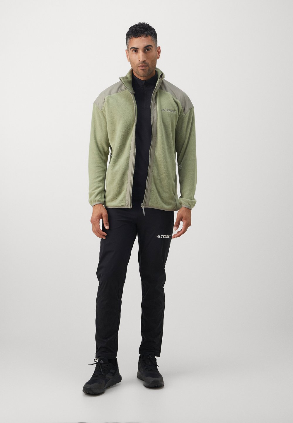 Grøn fleecejakke med høj krave, lynlås og grå detaljer, parret med sorte bukser. Teksturen er blød; jakken har et logo.