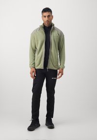 Grøn fleecejakke med høj krave, lynlås og grå detaljer, parret med sorte bukser. Teksturen er blød; jakken har et logo.