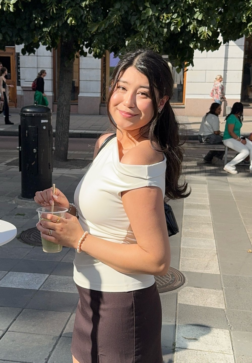 Vrouw die een doorzichtige beker met een rietje vasthoudt, gekleed in een aansluitend wit off-shoulder shirt en een donkere minirok, in een stedelijke buitenomgeving.