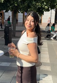 Vrouw die een doorzichtige beker met een rietje vasthoudt, gekleed in een aansluitend wit off-shoulder shirt en een donkere minirok, in een stedelijke buitenomgeving.