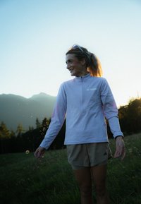 Lyse lilla bluse med lange ærmer, høj krave og lynlås, parret med beige shorts. Stoffet ser let og åndbart ud.