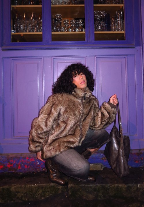 Veste en fausse fourrure dans des tons de marron, coupe oversized, portée avec un jean noir et des bottes en cuir marron. Tenant un sac à main en cuir noir.