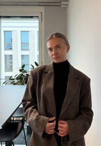 Hnedý vlnený blejzer so štruktúrovaným dizajnom, s klopami a veľkými rukávmi, nosený cez čierny rolák, v interiéri s rastlinou v pozadí.