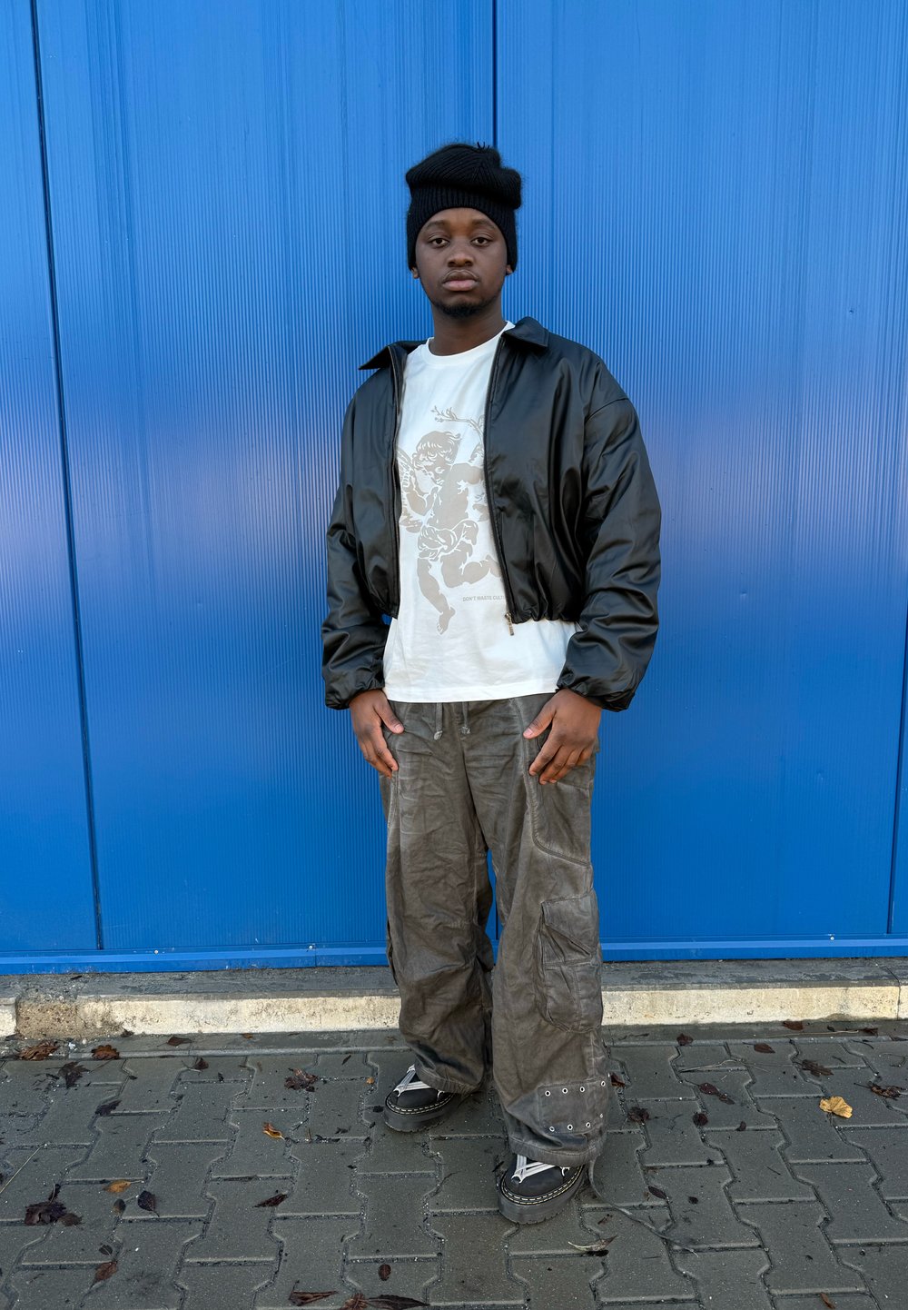 Fekete rövidített bomber dzseki, fehér grafikás póló, bő szürke cargo nadrág és fekete bakancs ezüst részletekkel, kék háttér előtt.