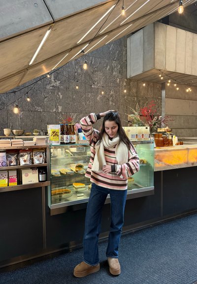 Mujer con suéter de rayas y bufanda está de pie dentro de una cafetería moderna cerca de una vitrina con pasteles y productos envasados, bajo luces colgantes de cuerda.