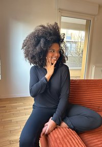 Una persona con un top nero a maniche lunghe e pantaloni neri è seduta su un divano arancione texture, con capelli ricci voluminose e una mano poggiata sul volto.