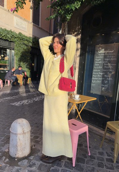 Suéter de punto amarillo y falda larga a juego, con un bolso de mano rosa texturizado. Pavimento de empedrado y un ambiente de cafetería al fondo.