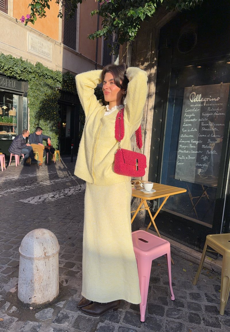 Yellow knitted sweater and matching long skirt, with a textured pink handbag. Cobblestone pavement and café setting in the background.