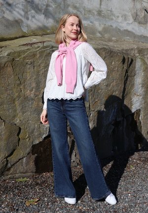 Mujer de pie al aire libre junto a una pared de roca, con una blusa blanca con ojales, vaqueros azules, zapatillas blancas y un suéter rosa sobre los hombros.
