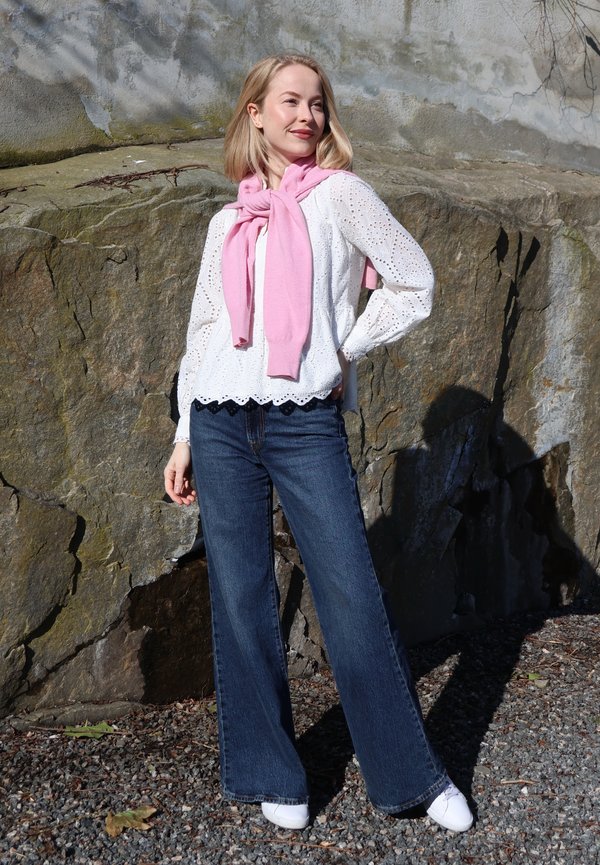 Žena stojí venku u skalní zdi, má na sobě bílou halenku s vyšíváním, modré džíny, bílé tenisky a růžový svetr přehozený přes ramena.