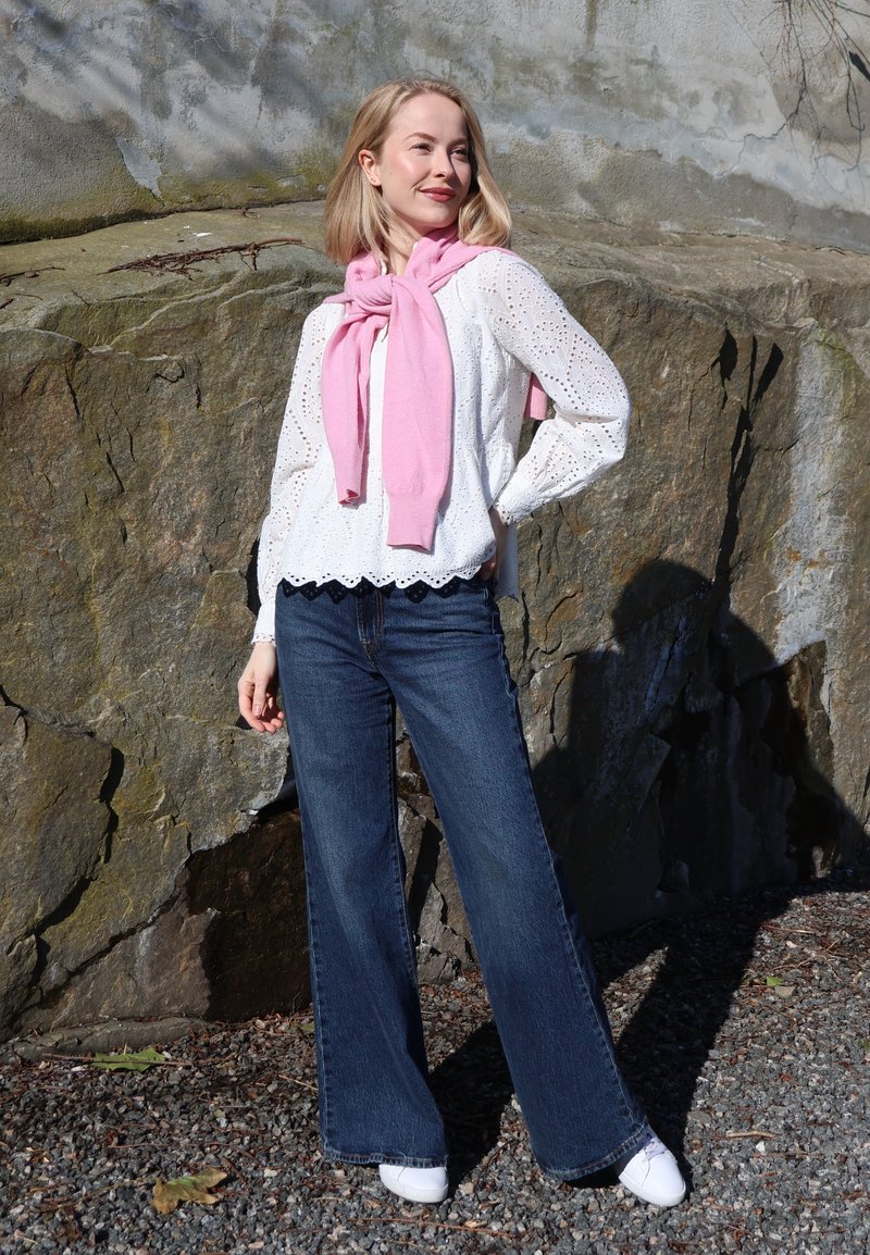 Femme debout à l'extérieur près d'un mur de rochers, portant un chemisier blanc à œillets, un jean bleu, des baskets blanches et un pull rose posé sur ses épaules.