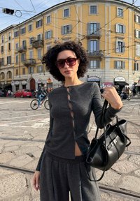 Maglione grigio a maglia con dettagli in bottone e aperture laterali, abbinato a pantaloni grigi coordinati. Borsa nera con hardware in argento. Ambientazione urbana.