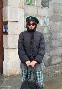 Personne portant un béret vert avec un logo blanc, une veste matelassée noire, des lunettes de soleil foncées, un pantalon à carreaux, debout contre un mur en pierre tenant un sac noir.