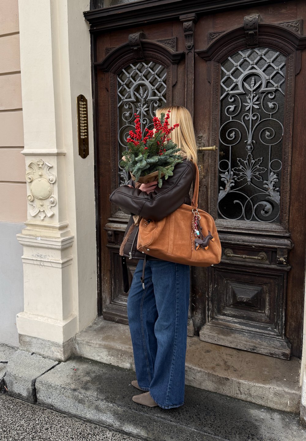 Femme aux cheveux blonds portant une veste sombre et un jean bleu, tenant des baies rouges festives et du feuillage devant une porte en bois ornée.