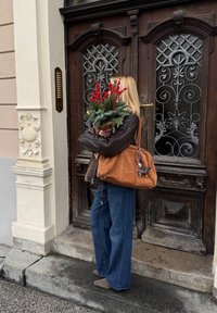 Femme aux cheveux blonds portant une veste sombre et un jean bleu, tenant des baies rouges festives et du feuillage devant une porte en bois ornée.
