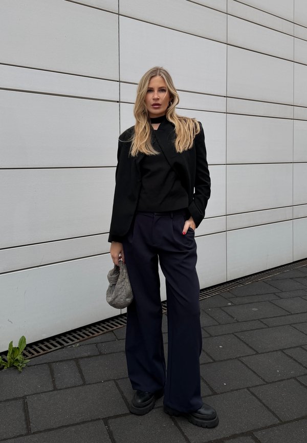 Vrouw met lang blond haar, een zwarte jas, een zwart topje, donkere wijde broek, zwarte schoenen, houdend een grijze gevlochten tas, staand op het trottoir nabij een witte muur.