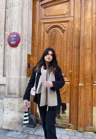 Una puerta de madera con detalles ornamentales; una persona sostiene una bolsa de compras a rayas y lleva un abrigo negro sobre una bufanda beige, de pie sobre adoquines.