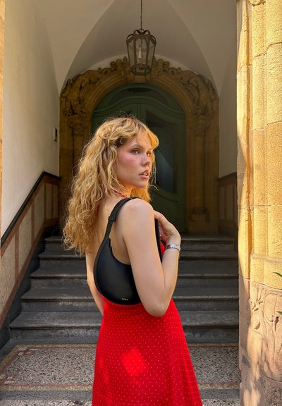 Bolso de hombro negro con diseño curvado y correa ajustable, combinado con un vestido rojo de lunares. El fondo presenta una arquitectura ornamentada.