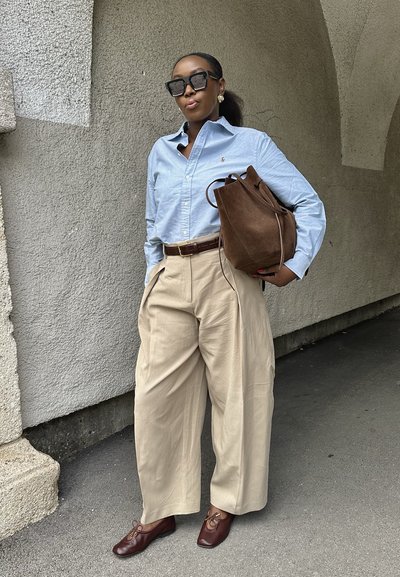 Mujer vestida con una camisa azul claro, pantalones anchos beige, zapatos marrones, un gran bolso marrón y gafas de sol negras, de pie contra una pared texturizada.