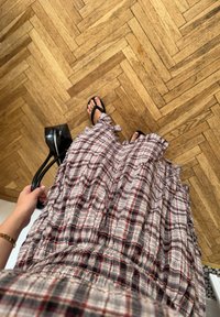 Rutete, volangskjørt i beige, rødt og grått stoff; svart håndveske i hånden og svarte flip-flops på føttene; parkettgulv i fiskebensmønster.