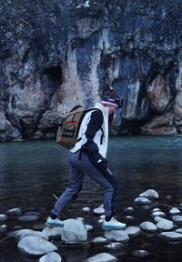 Personne traversant un ruisseau sur de grosses pierres, portant une veste noire et blanche, un pantalon gris et un sac à dos vert. Rochers enneigés en arrière-plan.