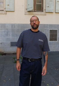 Mężczyzna w okularach, w niebiesko-paskowanej koszuli, z tatuażem na lewym przedramieniu, stoi przy kamiennej ścianie z otwartymi okiennymi żaluzjami.