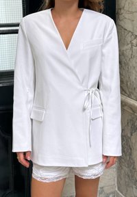 T-shirt enroulé blanc avec manches longues, fermeture à nouer à l'avant, et deux poches latérales. Tissu lisse avec un design épuré et un décolleté en V.