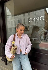 Gestreepte roze en witte cropped shirt met losse mouwen, gecombineerd met lichtblauwe wijdere jeans. Een geweven tas met een bloemaccent wordt vastgehouden.