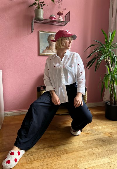 Camisa blanca de botones con estampado de corazones rojos, pantalones de pierna ancha en azul marino y zapatillas de casa extra suaves con puntos rojos. Entorno interior con paredes rosas y plantas.