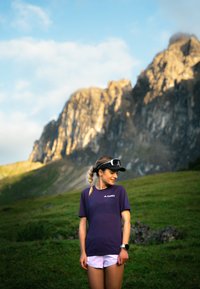 Lilla treningst-skjorte med korte ermer, kombinert med lyse shorts. Personen har en svart caps og solbriller, med en røff fjellbakgrunn.