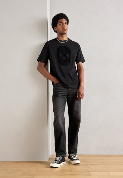 Camiseta negra con un gráfico de calavera, combinada con jeans oscuros y zapatillas en blanco y negro. El modelo está de pie contra un fondo neutro.