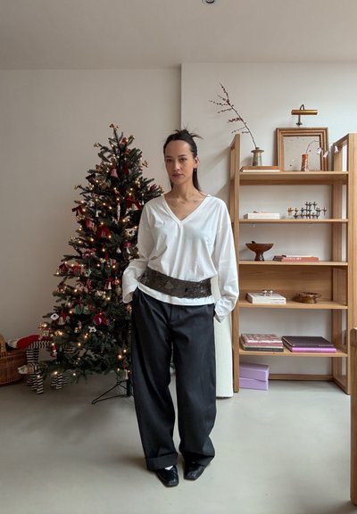 Top de manga larga blanco con escote en V, pantalones negros de pierna ancha y un cinturón negro texturizado. El fondo presenta un árbol de Navidad decorado.
