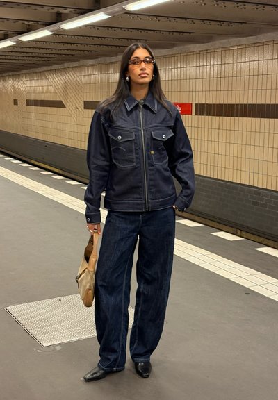 Chaqueta de mezclilla oscura con dos bolsillos en el pecho, combinada con jeans oscuros de pierna ancha. Zapatos negros de punta y un bolso marrón claro.