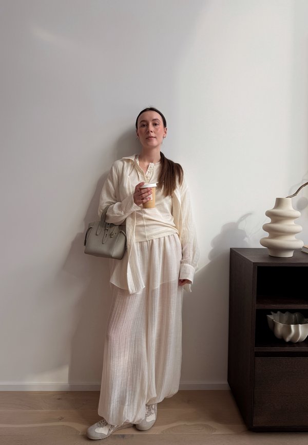 Vrouw in losse, lichtgekleurde kleding die een koffiekop vasthoudt, staand tegen een vlakke muur naast een donkere kast met een sculpturaal vaasje.