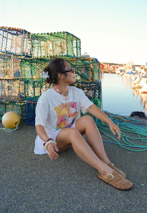 Femme assise sur un quai près de cages à homards empilées et de cordes vertes, portant un t-shirt blanc à motif graphique, un short, des sandales et des lunettes de soleil.