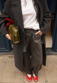 Manteau gris foncé surdimensionné, t-shirt blanc, pantalon gris plissé avec un bouton, et chaussures en cuir verni rouge. Sac vert avec un accent rouge.
