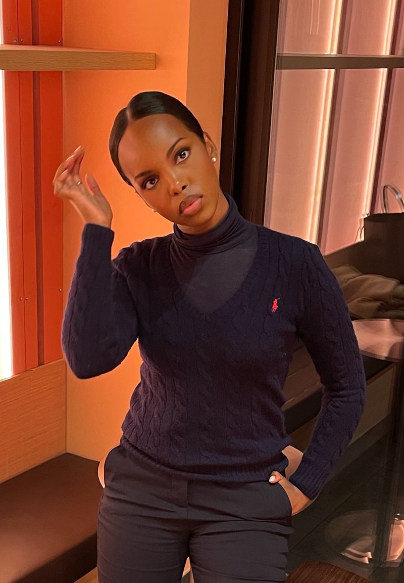 Maglione blu navy a maglia intrecciata con scollo a V e maniche lunghe, abbinato a pantaloni scuri. Vengono indossati orecchini semplici in argento.