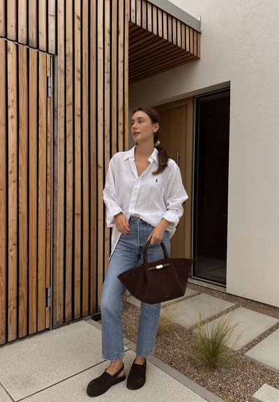 Camisa de lino blanca, jeans de mezclilla azul claro, bolsa de ante marrón oscuro y zapatos. De pie cerca de una pared de tablones de madera, con una puerta minimalista al fondo.