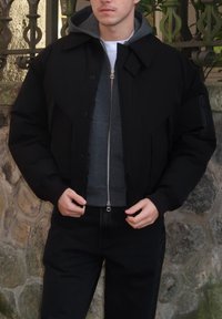 Veste bomber noire avec un design matelassé, dotée d'une fermeture éclair, d'un grand col et de poches avant. Portée par-dessus un sweat à capuche gris avec fermeture éclair et un jean foncé.