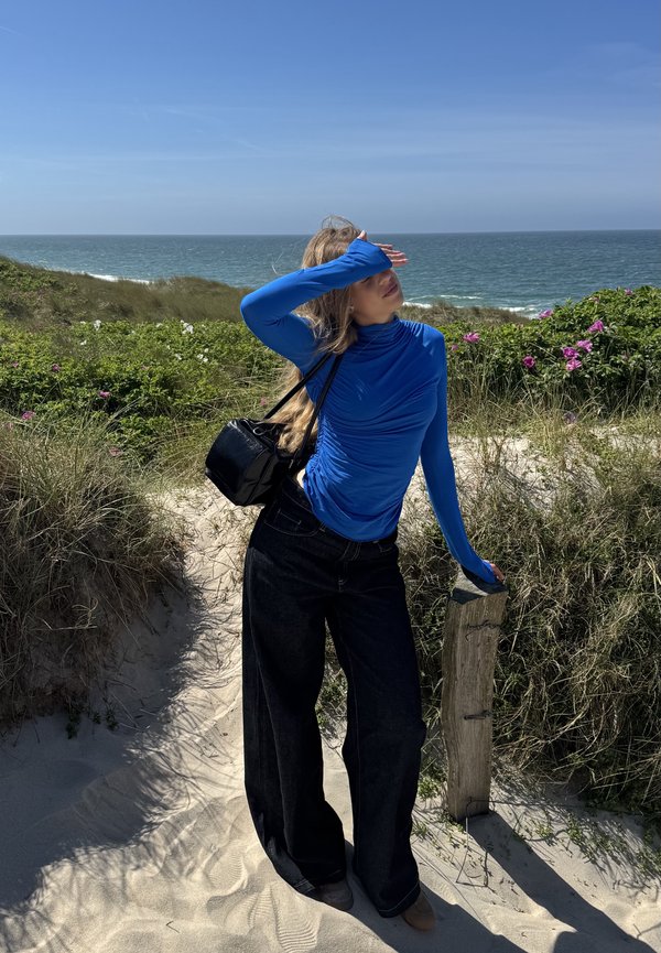 Femme en haut bleu et pantalon noir se tenant sur un chemin sablonneux près de l'océan, se protégeant les yeux, avec de la verdure et des fleurs derrière elle.