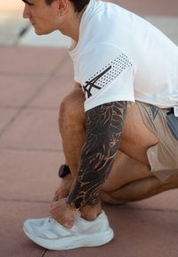 Atletisch figuur in een wit shirt met zwarte accenten knielt op roze trottoir, veters van witte sneakers met drie diagonale strepen strikkend. Gedetailleerde tatoeage op de arm.