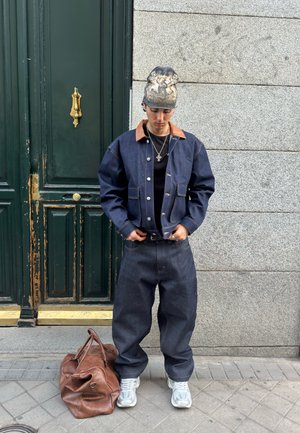 Joven con chaqueta y pantalones de mezclilla oscura, zapatillas blancas, gorro con estampado, ajustando el cinturón, de pie junto a una gran bolsa de cuero marrón en la acera.