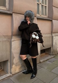 Svart kort blazer over et svart antrekk, knelengde shorts, høye svarte støvler og en grå pelslue. Holder en brun veske med hodetelefoner.